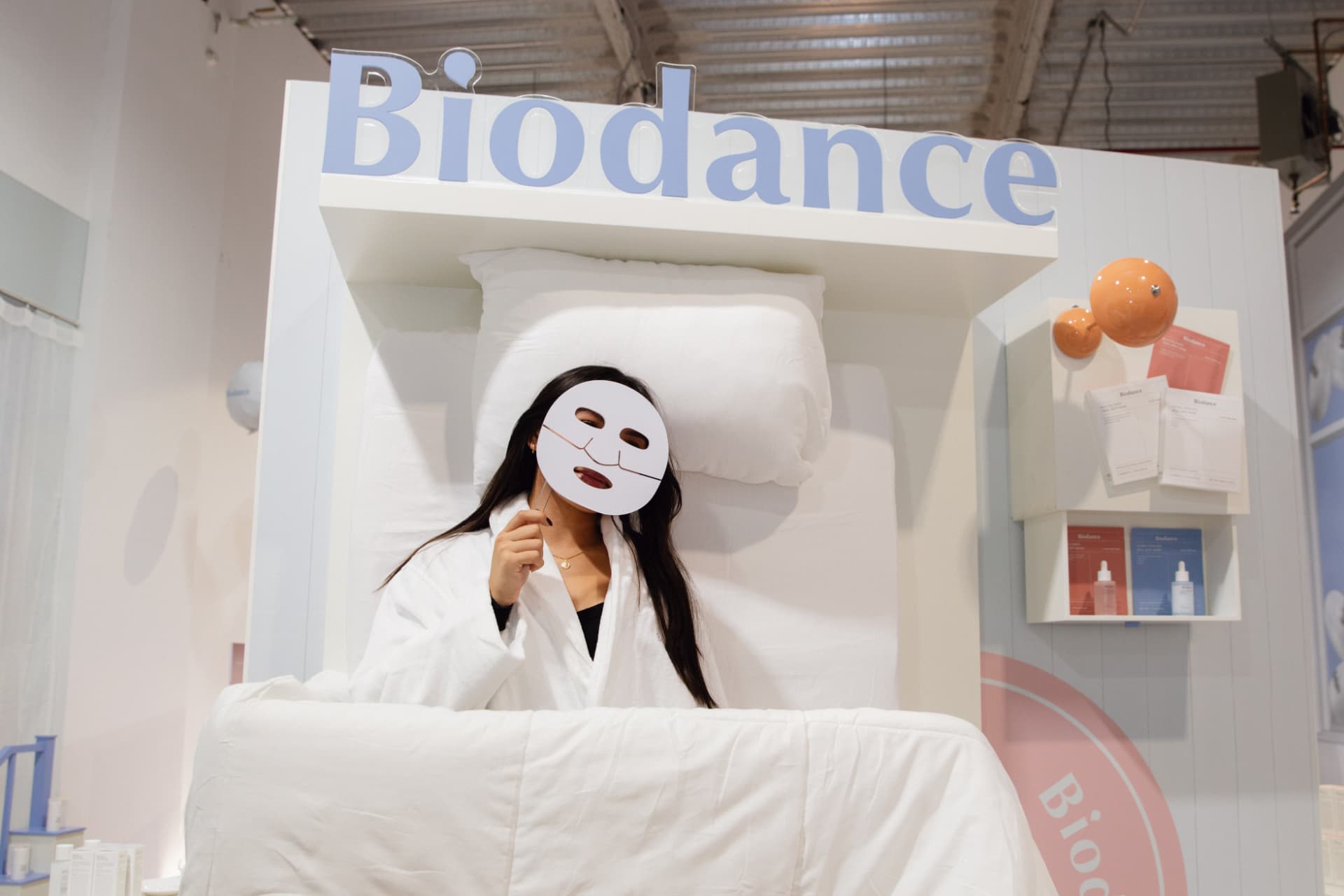 Guest in a robe holding a Biodance sheet mask in the branded bed photo set